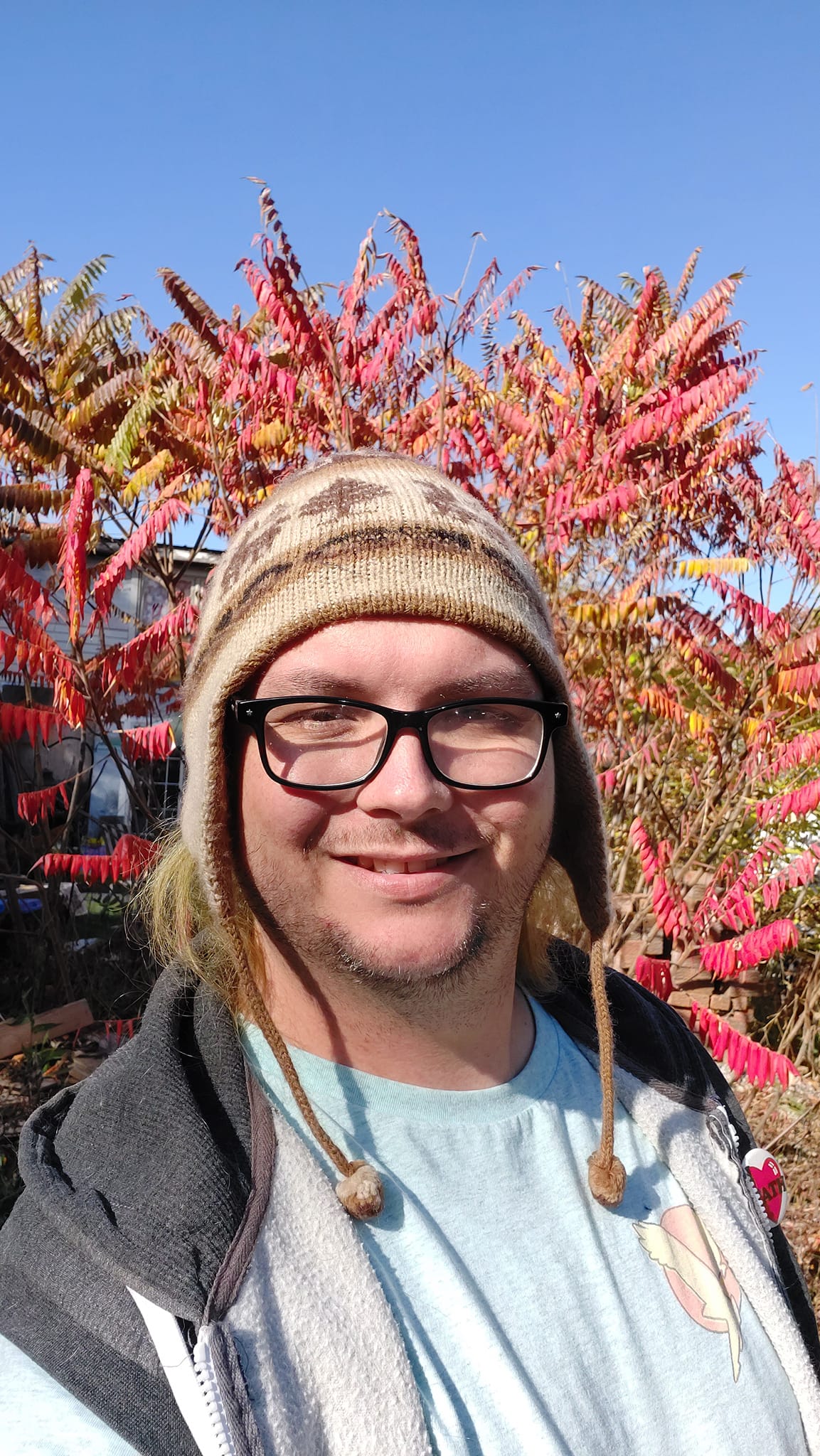 A man is wearing a toque, standing in front of bright red sumac leaves